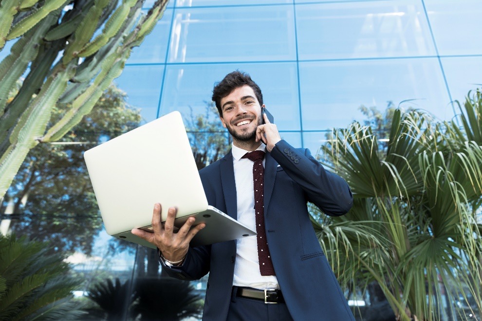 A person in business attire holding a laptop and talking on a phone in front of a modern glass building with surrounding greenery.