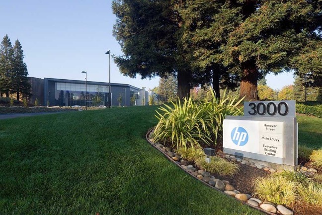 A modern building with a glass facade, surrounded by a green lawn and  trees. In the foreground is  a sign with “3000 Hanover Street, HP logo and Executive Briefing Center."